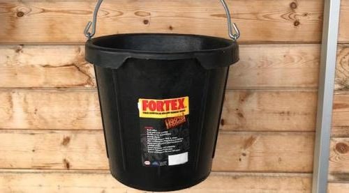 A black plastic Fortex bucket with a metal wire handle, hanging against a light brown wooden plank wall. The bucket features a prominent yellow and red Fortex logo label on the front.