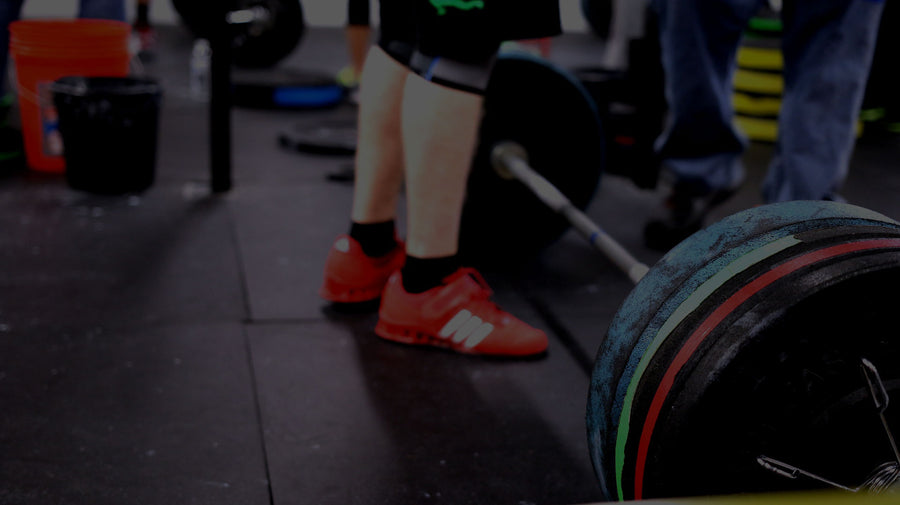 A close-up, low-angle view of a person in a gym lifting a barbell. The person is wearing black weightlifting shorts and bright red athletic shoes, standing on a dark rubber gym floor next to a loaded barbell with colorful weight plates.