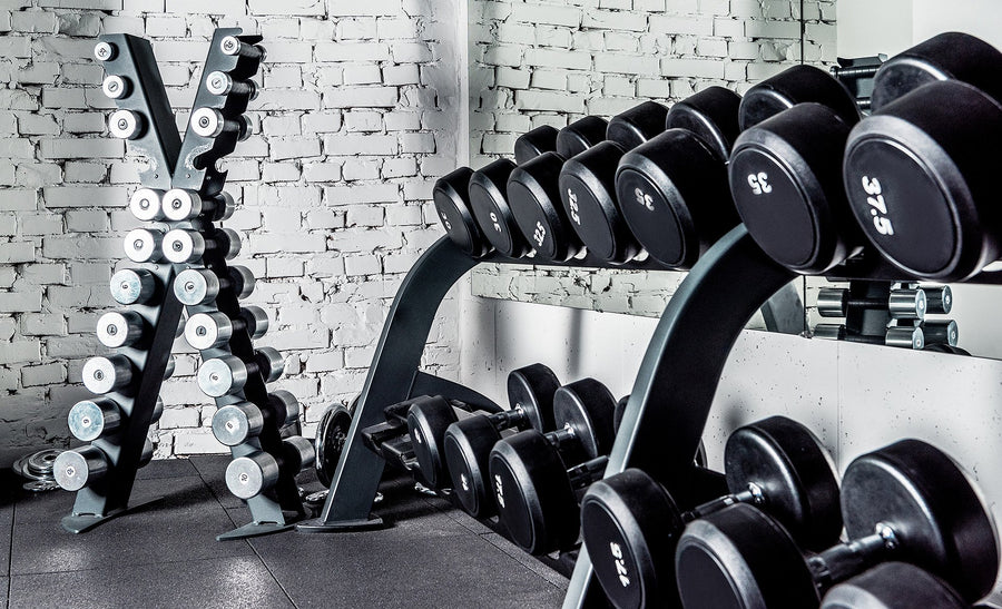 Black adjustable dumbbells arranged on a rack in a gym setting with a white brick wall background.