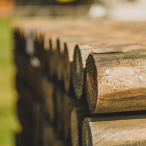 A close-up, shallow depth-of-field view of stacked natural wood logs or fence posts, with the front log in sharp focus and the background and sides blurred.