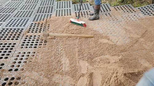 A person's boot and hand are visible while using a brush to spread a layer of light brown sand over dark gray interlocking drainage grid tiles, with a pickaxe lying nearby.